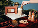 A styled holiday scene showing a boxed gingerbread chai drink mixer cube kit on a white pedestal next to a whipped latte topped with a gingerbread cookie, with a hand reaching in. Decorative gingerbread trees, wrapped gifts, a plate of cubed sweets, and a French press are arranged on a red draped surface against a blue backdrop with evergreen branches.