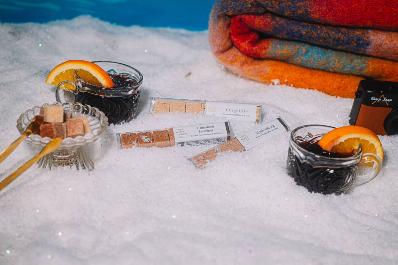 Two mulled wine glasse cups with orange slices, a LUXE Mixer Cube dish, and wrapped Luxe Mixer Cube Minis on a snowy surface.