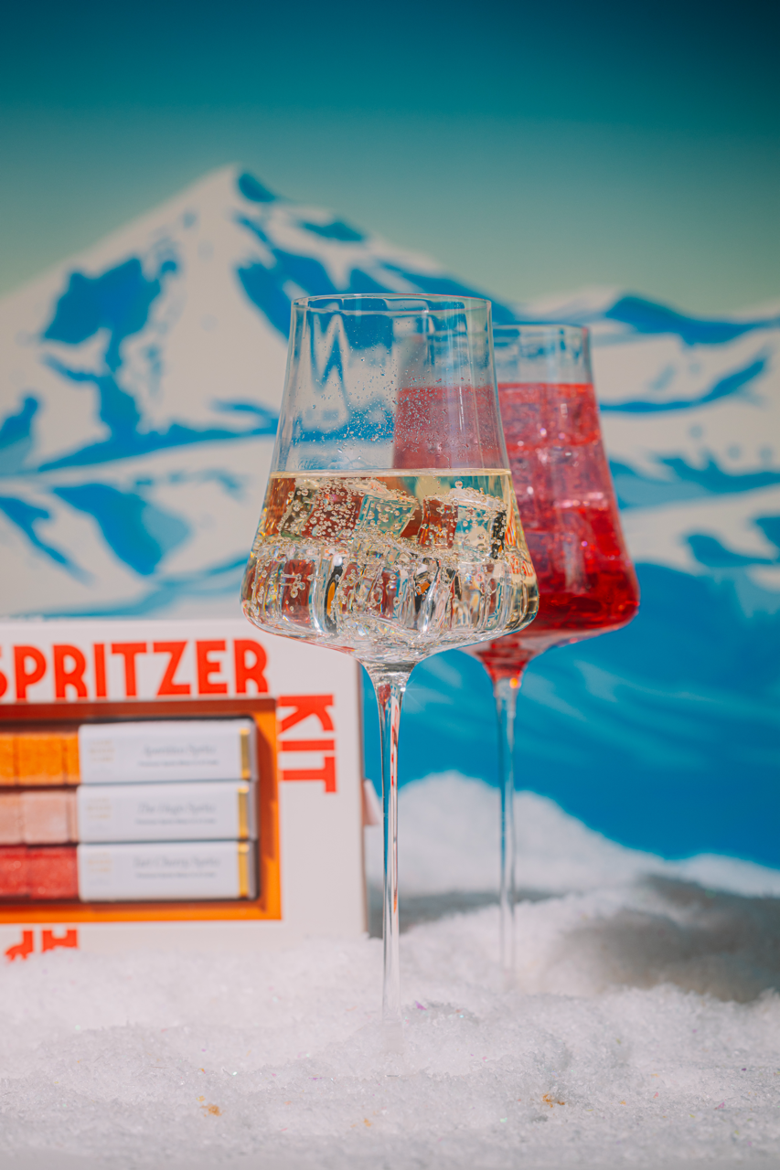 Two glasses of sparkling wine with a mountainous background and Spritzer Kit packaging.