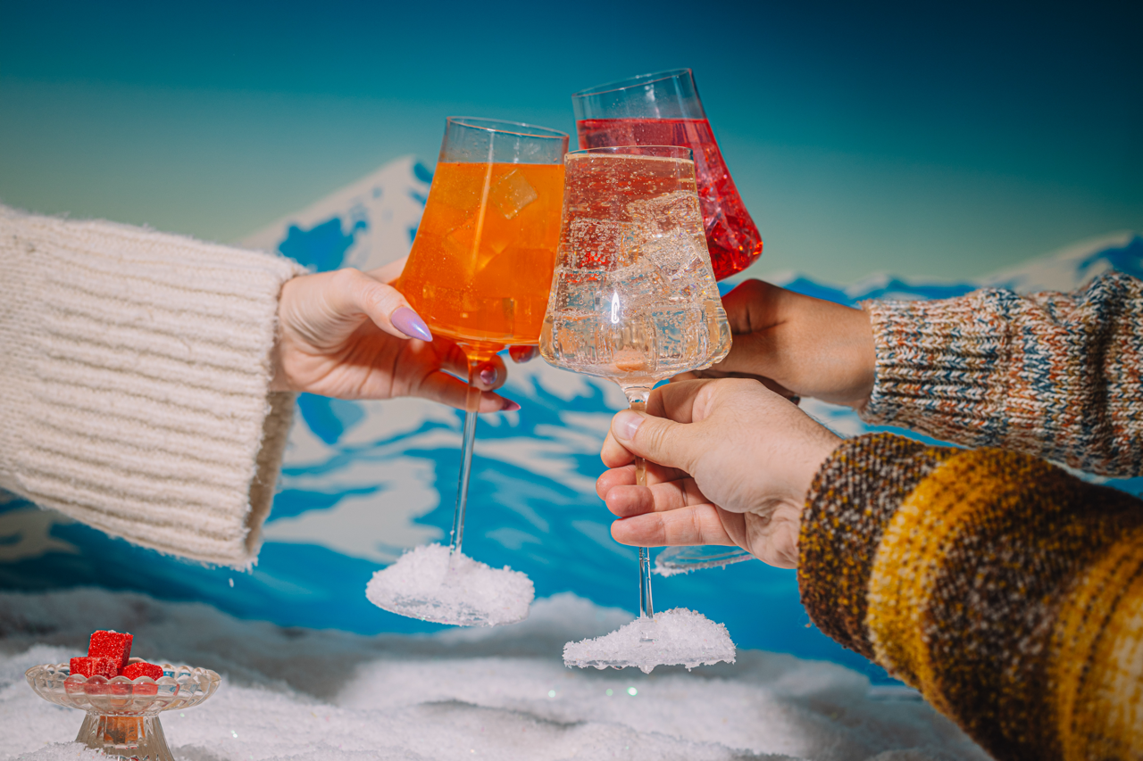 Thre people clinking glasses of spritzer kit drinks, against a snowy mountain background. 