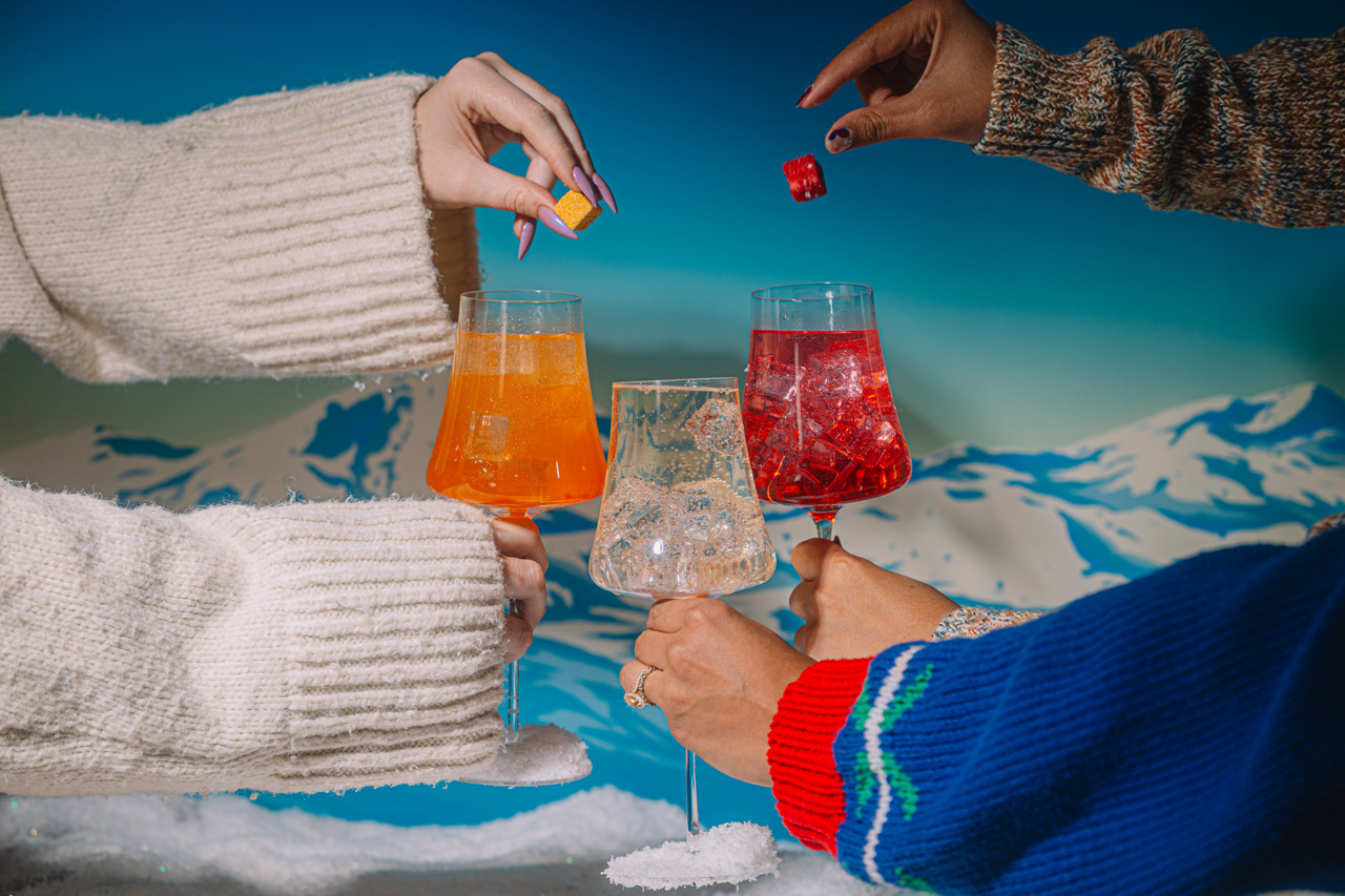 Three people holding glasses of colorful winter spritzers, left and right dropping Luxe Mixer Cubes into the spritzers.  against a snowy landscape.