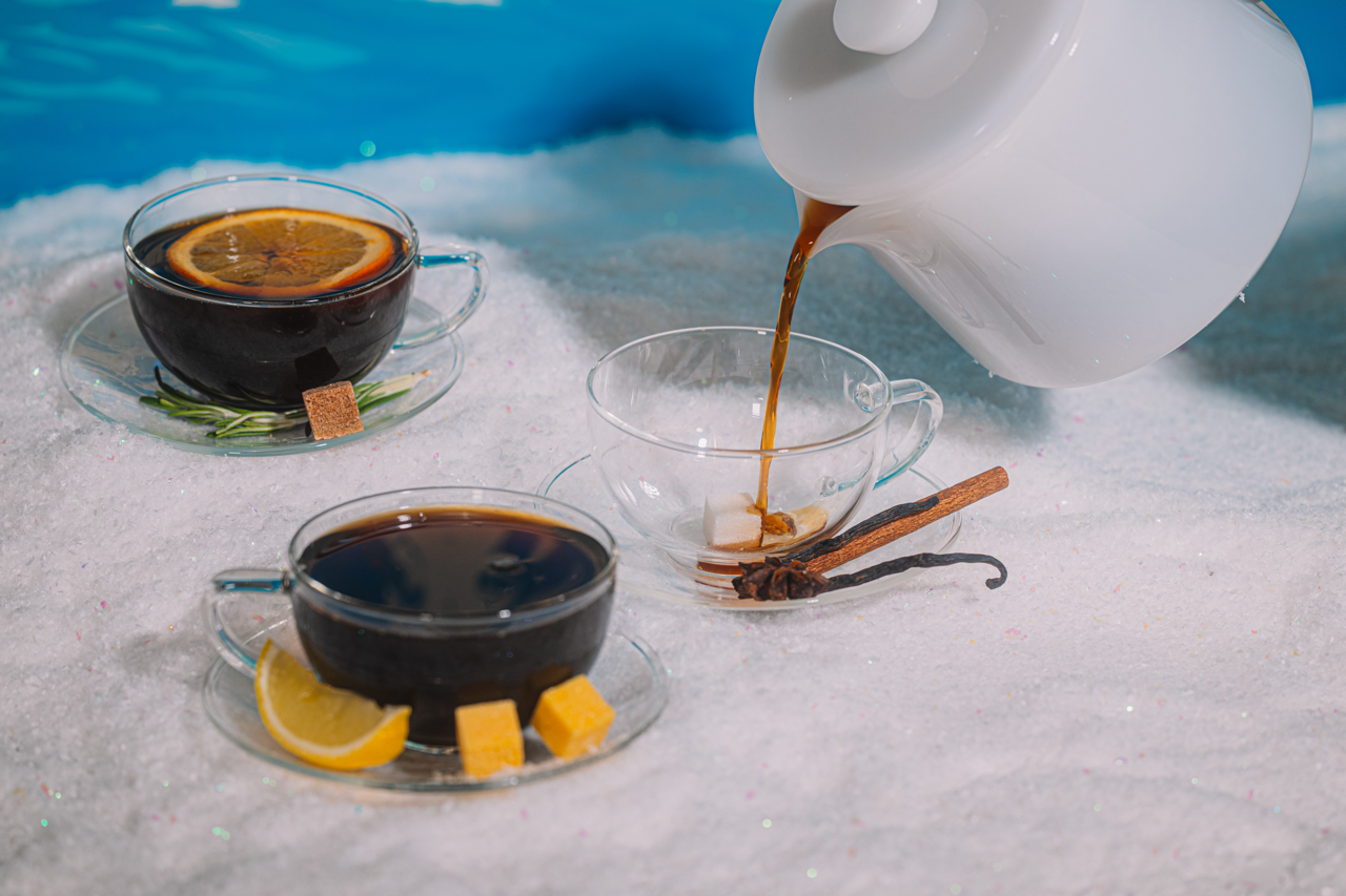 Tea being poured into a glass cup with a white teapot, surrounded by tea leaves and a vanilla bean on a white surface.