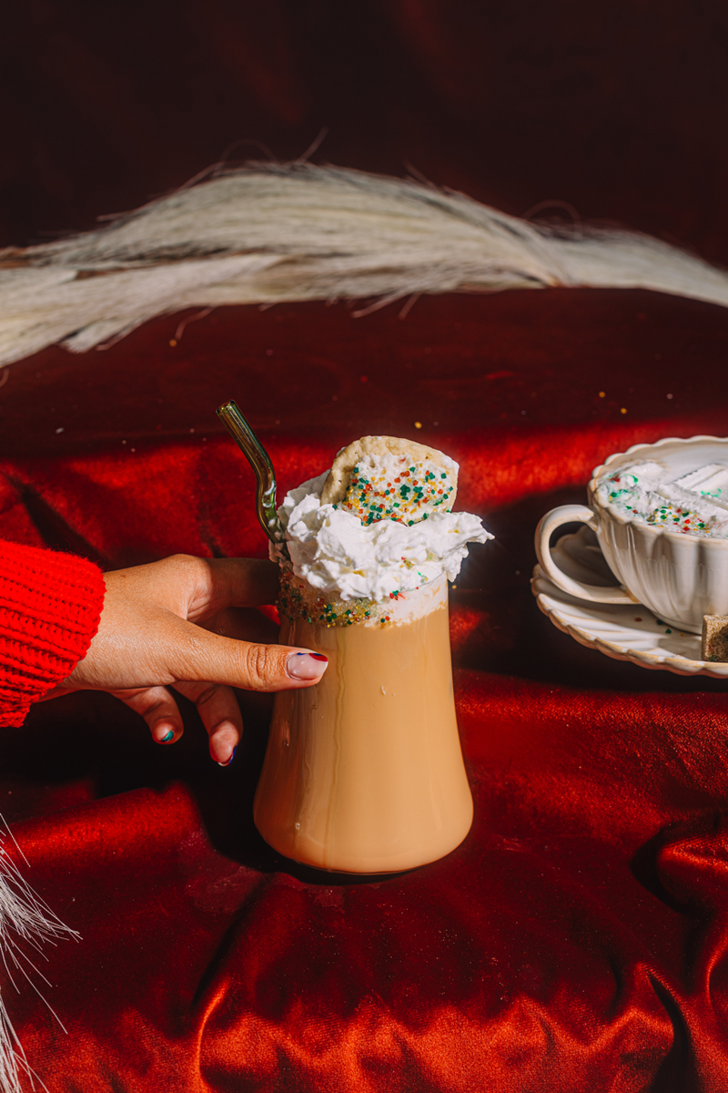 Hand holding a tall glass of iced coffee with whipped cream and sprinkles, on a red surface.