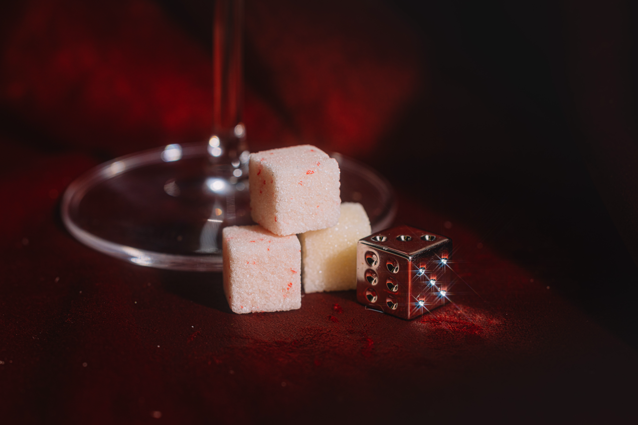 Stacked LUXE Mixer Cubes and a Prada silver dice on a dark red velvet surface with a blurred martini glass in the background.