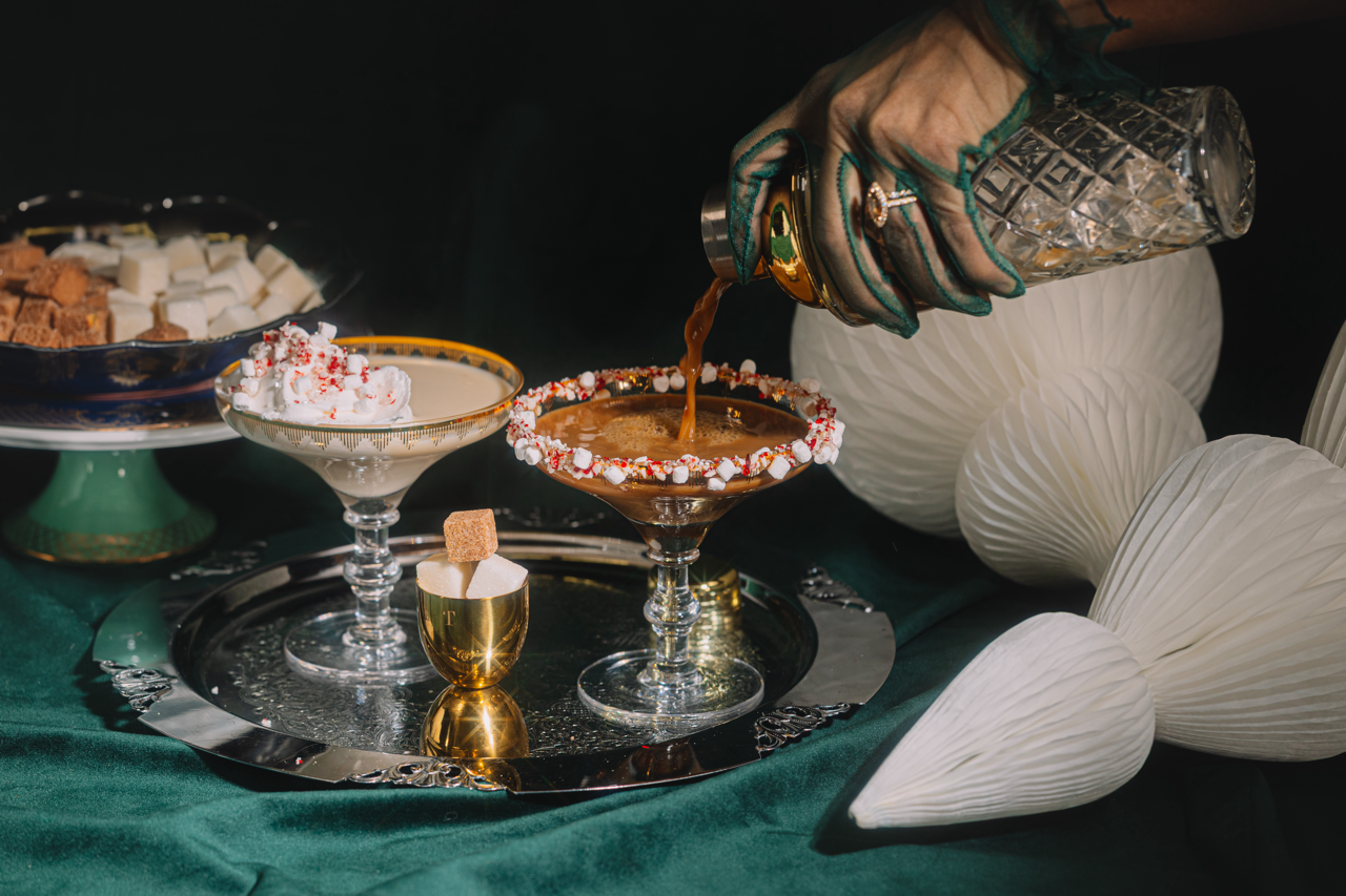 Person pouring a drink into a cocktail glass on a dark surface with decorative elements.