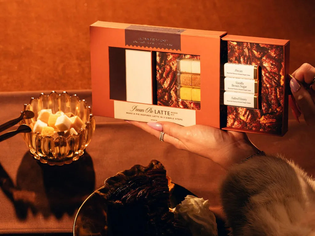 A hand holds an open box of Pecan Pie Latte sugar cubes against a warm, cozy background with a glass bowl of  with an assortment of sugar cubes and a plate of pecan  pie.