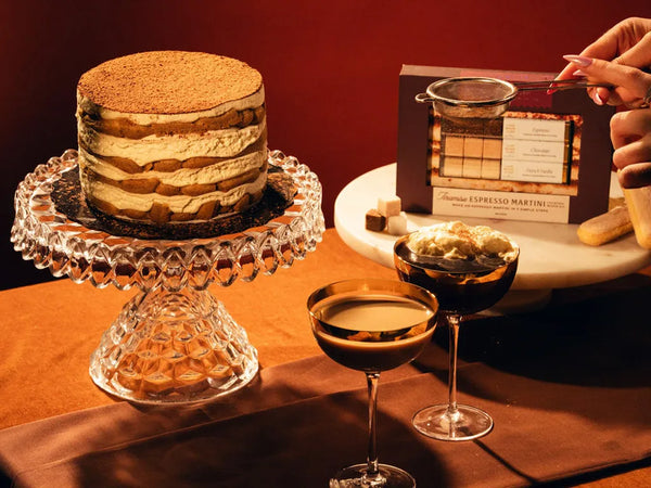 A layered dessert cake on a crystal pedestal stand, with two coupe glasses filled with espresso martini, and a boxed Tiramisu espresso martini cocktail mixer kit in the background. A hand is visible dusting the martini. 