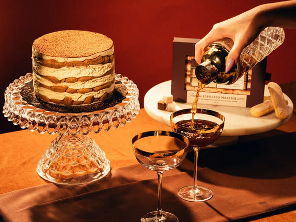 A hand pours a dark amber liquid from a bottle into two coupe glasses, with a layered dessert on a crystal pedestal and a Tiramisu espresso martini box in the background against a warm red-brown backdrop.