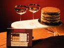 A boxed tiramisu espresso martini cocktail mixer kit sits on a table in front of two gold-rimmed coupe glasses and a layered dessert on a white pedestal, against a warm red-brown background.
