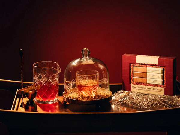 A styled product display featuring a box of Luxe Mixer Cube Maple Crumble Old Fashioned mixer cubes, a crystal mixing glass with red liquid, a crystal decanter, and a glass cloche on a dark wooden tray against a deep red background.