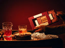 A hand holds a red box of gourmet old fashioned mixer cubes over a dark table styled with glasses of amber liquor, a crystal pitcher, and a silver tray, under warm red ambient lighting.