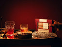 A hand holds a red gift box labeled 'Maple Crumble Old Fashioned' beside a bar cart with a cut-crystal pitcher, a glass of amber liquid, and a crystal trivet on a dark wooden table against a deep red background.