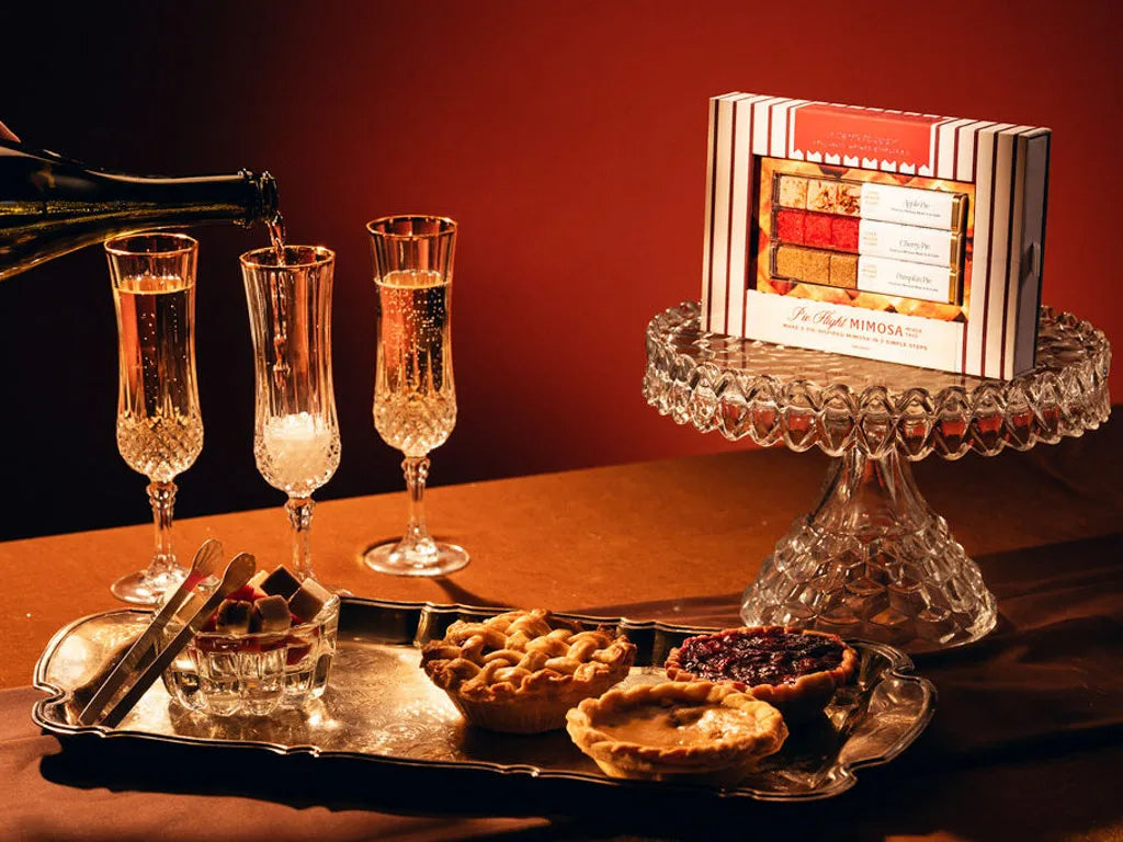 A boxed gift set labeled 'Pie Flight Mimosa' sits on a crystal pedestal against a warm red-brown background, with a bottle pouring into three champagne flutes and small assorted pies on a silver tray in the foreground.