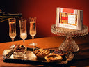 A boxed gift set labeled 'Pie Flight Mimosa' sits on a crystal pedestal against a warm red-brown background, with a bottle pouring into three champagne flutes and small assorted pies on a silver tray in the foreground.