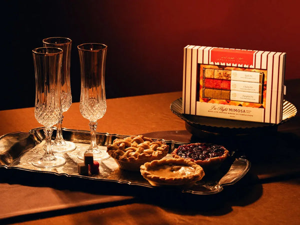 A boxed gift set labeled 'Pie Flight Mimosa' sits on a dark table beside a silver tray holding two champagne flutes, two small pies, and a couple of chocolates. The scene is lit with warm amber light against a dark background.
