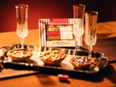 A Mimosa Cube gift set from Teaspressa displayed on a silver tray with three champagne flutes. The set includes an assortment of chocolate tartlets and the branded box, all photographed on a wooden table under warm, moody lighting.