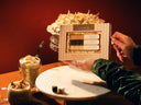 A person holds a boxed set of coconut cream matcha chocolates on a table, with a bowl of shredded coconut, two iced coffee drinks, and a small matcha cube on a white platter against a warm dark red background.