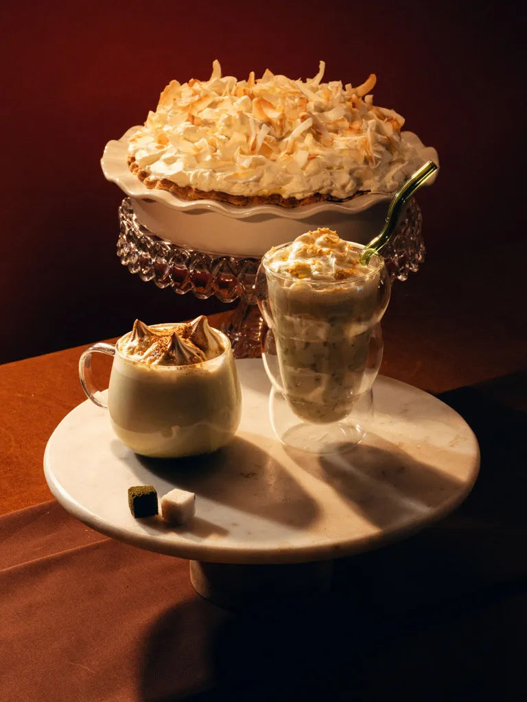 A dessert pie topped with whipped cream and coconut on a pedestal stand, alongside two glasses of whipped cream and caramel drizzle on a marble serving board, set against a dark warm brown background.