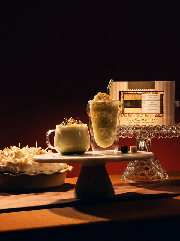 Two frothy dessert drinks displayed on a white pedestal stand, with a bowl of whipped topping and a branded card in the background against a dark warm-toned setting.
