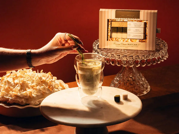 A hand holds a green tea leaf above a glass of matcha latte, with a box of Forest Chase Matcha on a crystal pedestal and a bowl of shredded coconut in the background against a dark red wall.